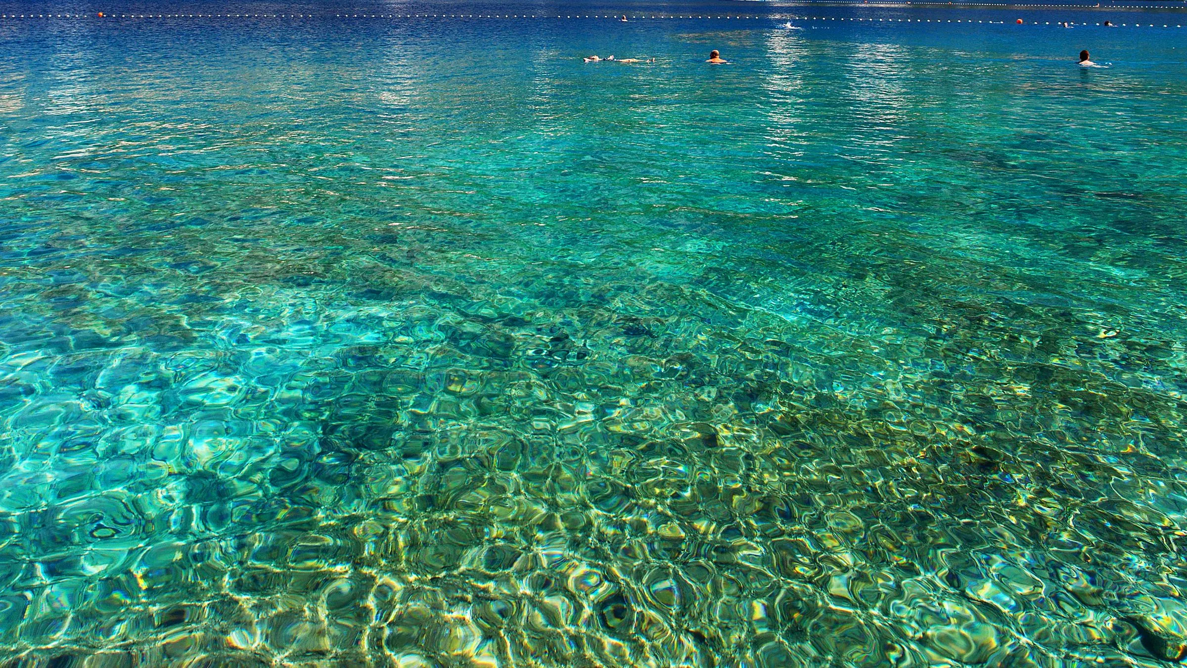 Clear turquoise Adriatic water shading from shallow to deep, with swimmers offshore