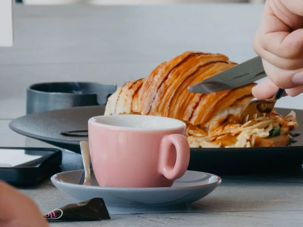 A cup of coffee beside a croissant being cut, on a table at Central Beach Split